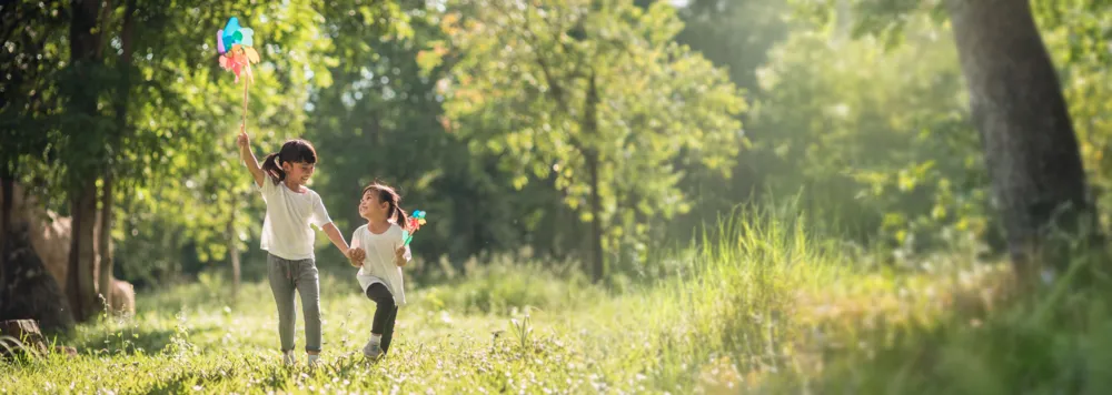 Two young girls holding hands and playing with colorful pinwheels in a sunlit grassy field