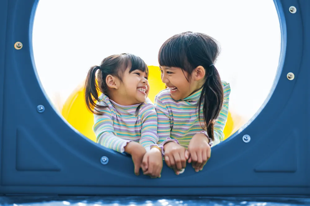 Two young girls smiling and leaning on the edge of a blue circular playground structure
