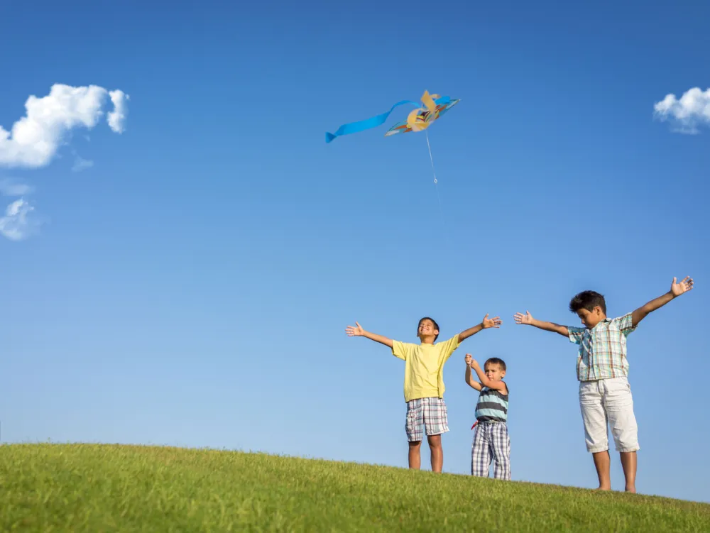Three children flying a colorful kite on a grassy hill under a clear blue sky
